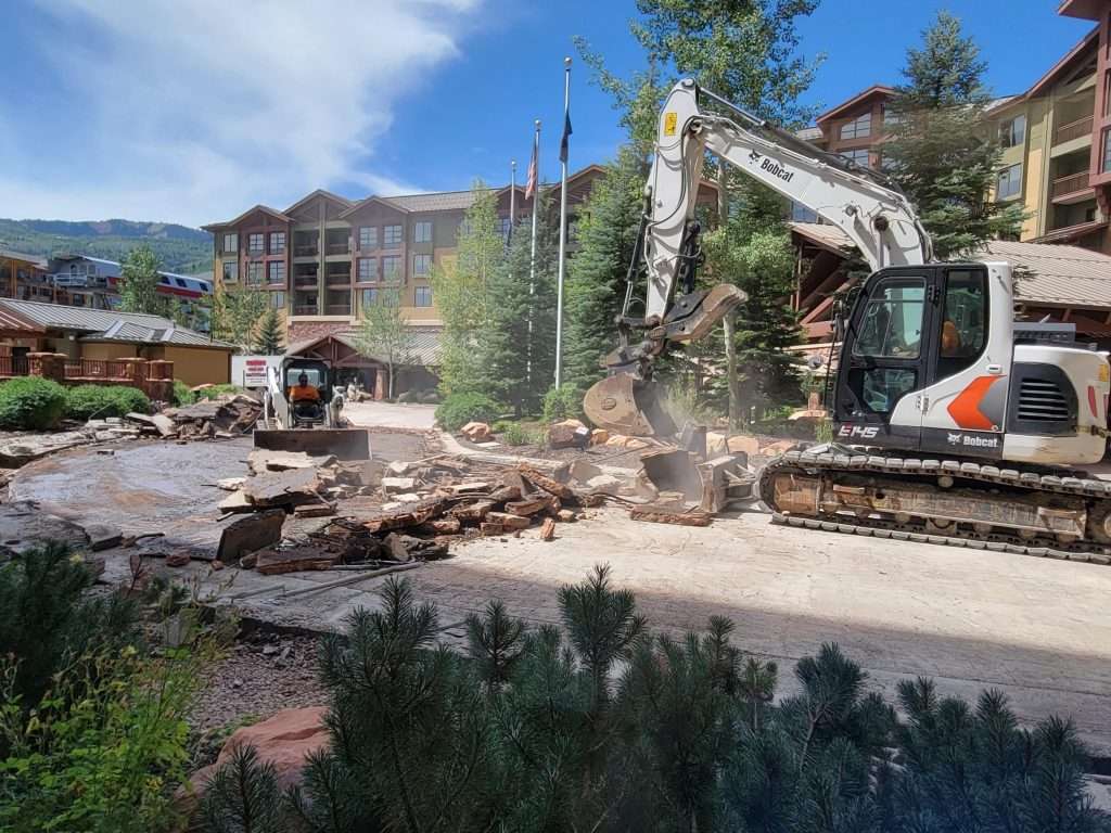 Excavator demolishing a building near a residential area on a sunny day.