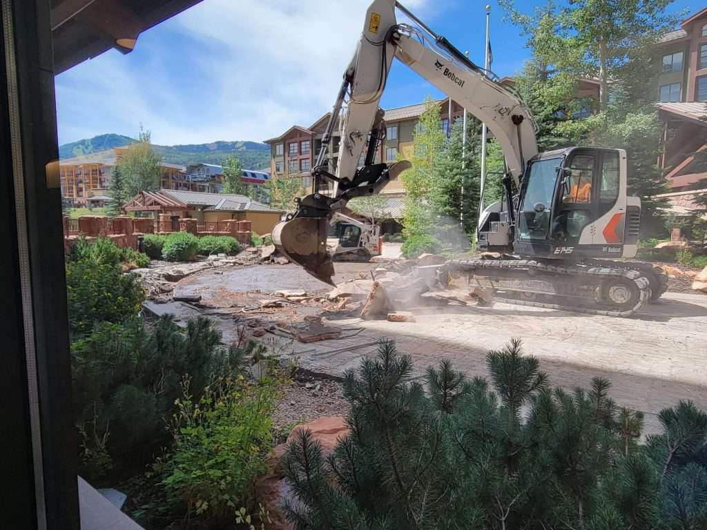 Excavator demolishing a building amid greenery and clear skies.