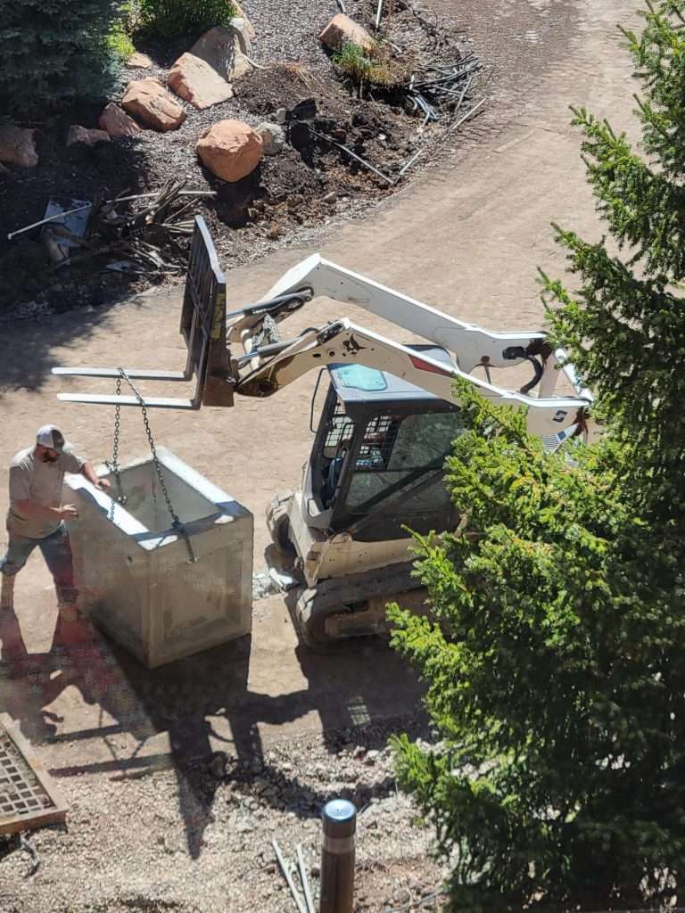 Workers operate a small excavator and a dumpster on a construction site.