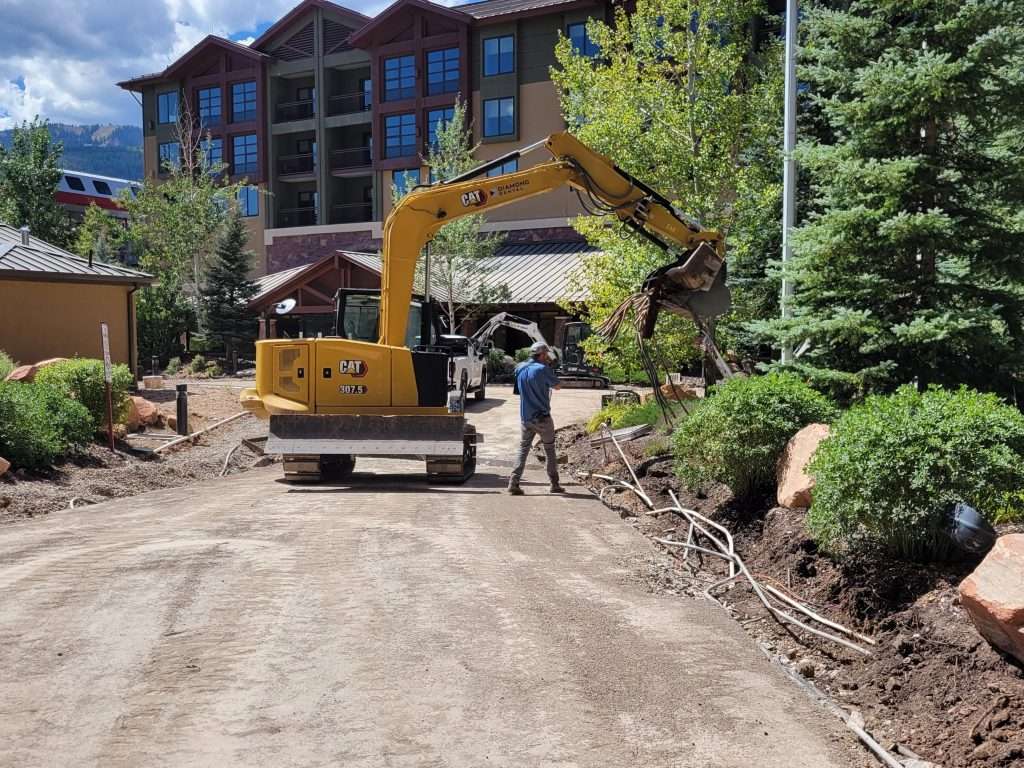 Excavator clearing a path near buildings and trees.