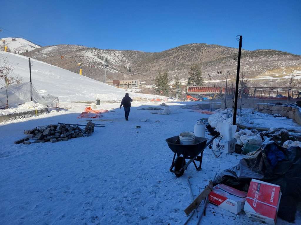 Person walking on a snowy path near a wheelbarrow with mountains in the background.