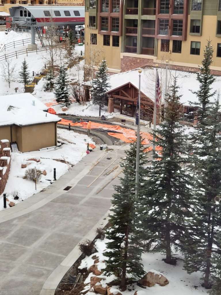 Snow-covered walkway with pine trees and buildings in the background.